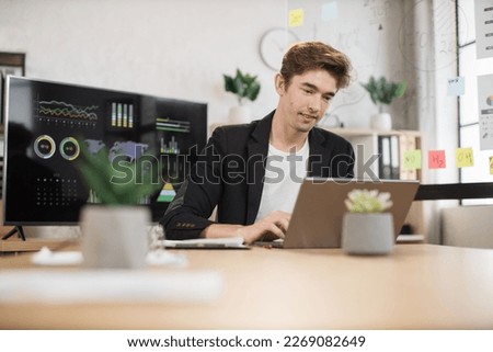 Confident caucasian man in white shirt sitting at office and using wireless laptop. Big monitor with various charts and graphs on background. Business and finance concept.