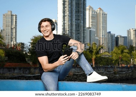 Young guy in a natural environment of the city while enjoying his music with headphones and his tablet.