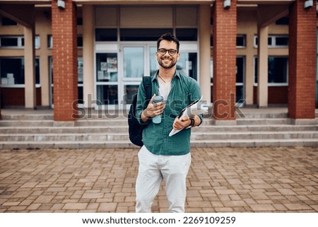 Happy male student standing in front of university building and looking at camera. Copy space. 