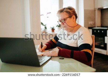 An elderly woman is pictured at home, using her laptop for e-learning purposes. She is wearing her glasses and focused on learning new information, age is just a number when it comes to education.