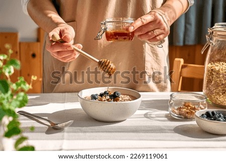 Woman preparing healthy dieting vegan nutritious breakfast. Female hand pouring honey in the bowl with oatmeal porridge with walnuts and blueberries. Royalty-Free Stock Photo #2269119061