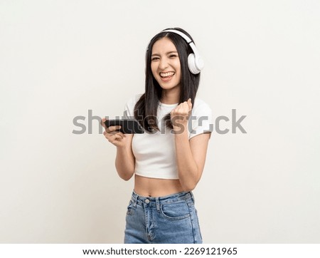 Beautiful young asian women play mobile game and put on wireless headphone standing on isolated background. Playing game on smartphone winning victory moment. Very enjoy and fun relax time