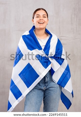 Happy young woman in casual clothes posing and smiling, wrapping herself in Greek flag against gray wall