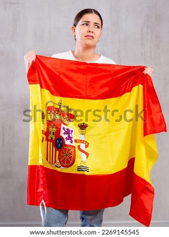 Sad young woman with Spain flag in hands posing sorrowfully against light unicoloured background