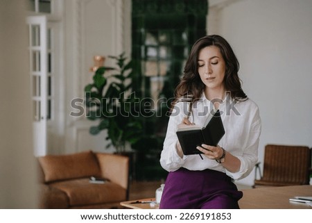 Overloaded brunette young woman in white shirt and violet pants sits on table writes in diary with tired face. Exhausted female holds diary against blurry interior. Businesswoman remote working.