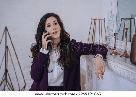 Puzzled brunetitehispanic young businesswoman in burgundy suit talks by phone leans on fireplace at home looks at camera. Gorgeous caucasian woman discussing with psychologist. Financial people.