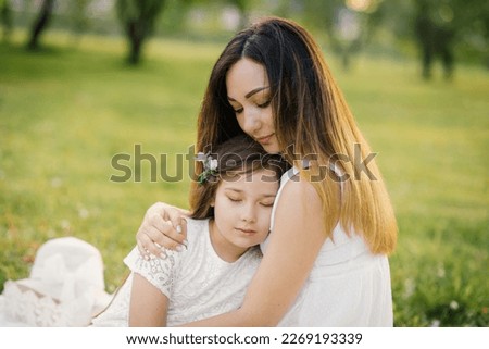 Mom hugs her daughter, sitting on a blanket in a spring blooming garden. Maternal love for a child