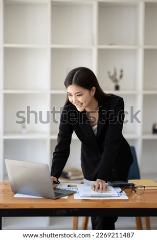 Young beautiful Asian businesswoman standing at her office desk while typing on laptop computer, working on her project before meeting.