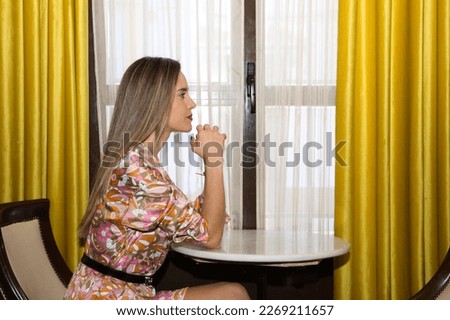 young and beautiful blonde woman sitting in an armchair next to a window covered with a curtain and yellow curtain. The woman is leaning with her elbows on the table and her hands clasped.