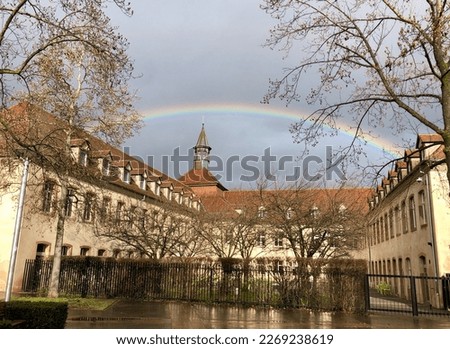 A colourful rainbow in a gloomy grey blue lilac sky just after rain. The rainbow forms an arch over an old European building with a gabled roof and a tower. Bare tree branches frame the picture