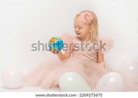 Beautiful girl in lush dress and hoop crown enthusiastically looks at small globe hands. White balloons isolated on white background.