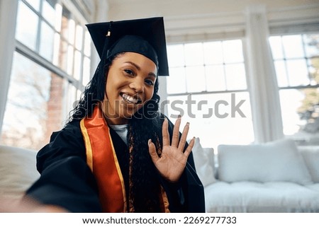 Happy African American woman in graduation gown taking selfie at home.