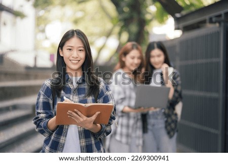 Asian college student focusing on laptop work or reading while other classmates in the background, outdoor portrait on campus campus.