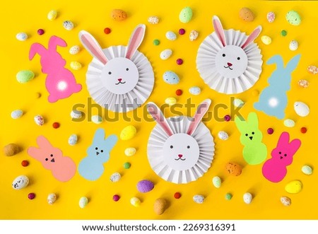 Craft, on a yellow background, cute, cute white paper rabbits and multi-colored Easter eggs.  Flat lay, top view.  Pattern, background picture, concept holiday light Easter.