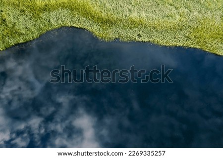 Aerial view on a river bank with green grass field and cloudy sky reflection.