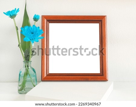 Brown wooden square photo frame blue flowers (chrysanthemum) in vase on the shelf have a white wall backdrop. 
