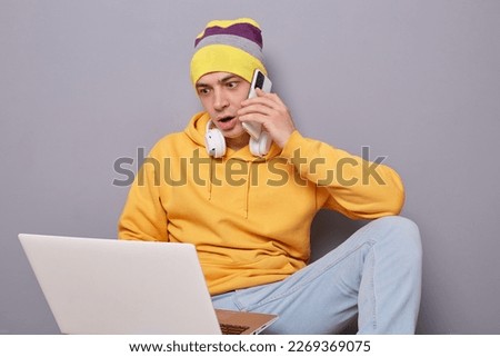Portrait of shocked man wearing beanie hat, yellow hoodie and jeans posing against gray wall, looking at laptop monitor with big eyes, talking on cell phone with somebody. Royalty-Free Stock Photo #2269369075