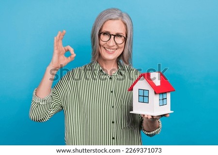 Photo of charming grandmother demonstrate okey symbol hold small house isolated on blue color background