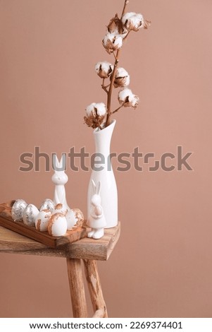 several branches with white cotton plant in white vase, golden easter eggs and rabbits - spring decoration on beige colored background,close up, negative space
