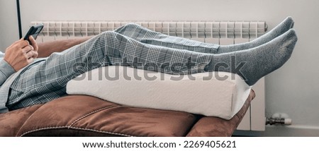 a man leans his legs on a leg elevation pillow made of memory foam, resting on the sofa, in a panoramic format to use as web banner or header Royalty-Free Stock Photo #2269405621