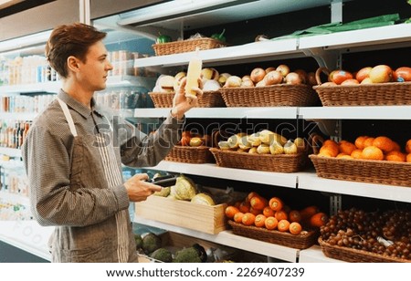 Caucasian young adult employee is working on checking the stock of fruit on shelves with barcode scanner. Salesman is holding a barcode scanner to examine the stock of corns in the basket on shelf.