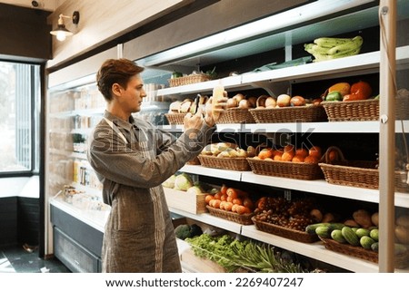 Caucasian young adult employee is holding corn and taking a picture with his smartphone to send to stock manager for quality checking. Salesman is checking the stock of fruit before opening the store.