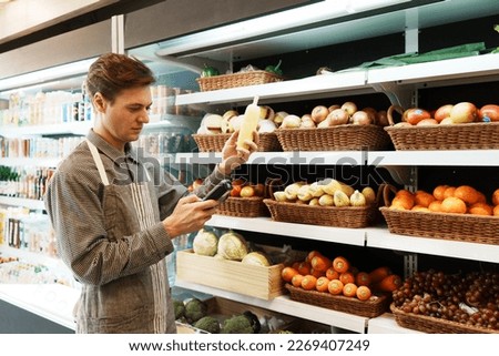 Caucasian young adult employee is filling out information about the quality and freshness of corn on the shelf with barcode scanner. Salesman is checking the stock of fruit in the basket on shelf.