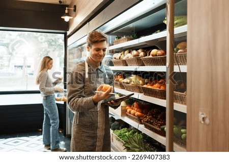 Caucasian young adult employee with an apron is checking quality of fruit on the shelves. Salesman is holding a tablet to record information about the quality and freshness of fruit in grocery store.