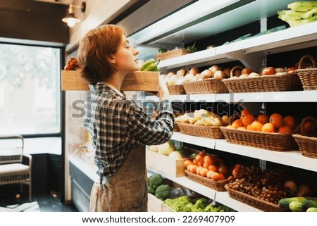Caucasian young adult saleswoman is carrying the basket of fruit and looking at shelves to add fruit until basket is fully. Female employee is sorting types of fruit before opening the grocery store.