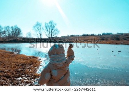 little child in yellow coat with white spots throwing stones into frozen lake