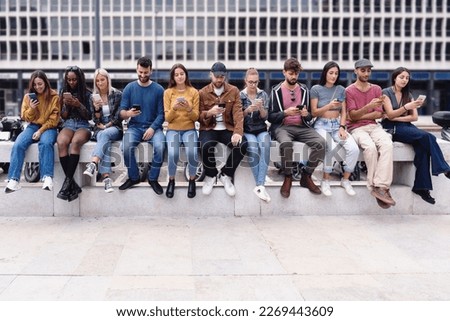 A group of eleven teenagers sitting on a wall, all with their heads down looking at their smartphones. The photo captures the concept of phubbing and social media addiction Royalty-Free Stock Photo #2269443609