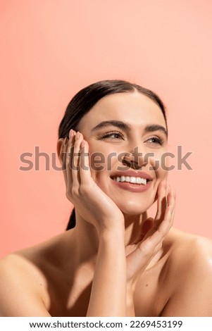 smiling woman with bare shoulders looking away while touching cheek isolated on pink