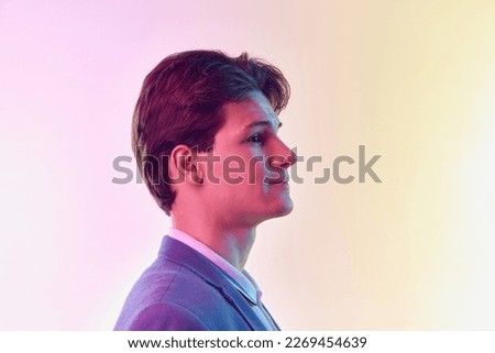 Profile view of young man, student graduate in jacket posing isolated over light gradient yellow-purple background in neon light. Emotions, fashion, art, education, business and ad concept.