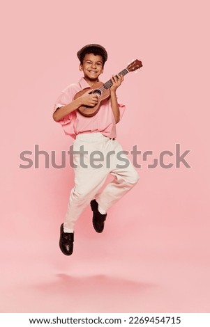 Happy emotional little african boy in retro style clothes and cap posing with ukulele guitar isolated on pink background. Concept of music, childhood, education, fashion and aspiration Royalty-Free Stock Photo #2269454715