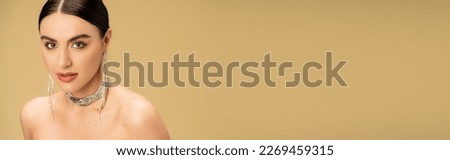 brunette young woman in necklace and earrings looking at camera isolated on beige, banner