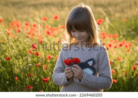 A little fair-haired girl stands in a field with wheat and red poppies in the summer.