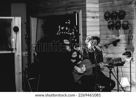 Young rocker musician with messy hair playing a wood electro acoustic guitar and singing in a music studio while him is recorded in video (in black and white)