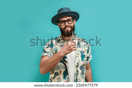 Portrait of a cheerful, joyful, stylish barber with a beard dressed in a shirt, hat, and glasses with scissors and a comb in his hands, looking at the camera, isolated on a blue background