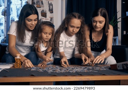  Staged photo. Lesbian couple and their kids are having a good time at home. Cozy atmosphere. Parents and girls sitting at the table with a lot of puzzles.                              