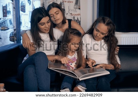   Staged photo. Lesbian couple and their kids are having a good time at home.  Parents and girls nestled on the couch. All are looking at a large illustrated book.                             