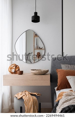 Modern, irregular shaped mirror above simple, wooden dressing table next to cozy bed in bedroom Royalty-Free Stock Photo #2269494461