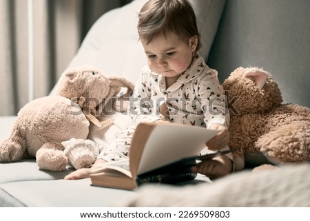 A little baby sitting on the sofa reading book. Teddy bear and rabbit toys are nearby