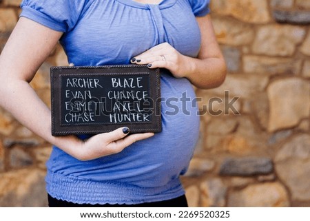 Pregnant woman standing in front of a rock wall holding a chalkboard sign with ideas for baby names