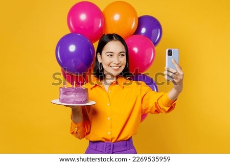 Happy fun young woman wear casual clothes celebrate near balloons hold cake with candles doing selfie shot on mobile cell phone isolated on plain yellow background. Birthday 8 14 holiday party concept