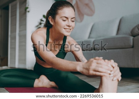 Full body close up young strong athletic fitness trainer instructor woman wearing green tracksuit train doing stretch lunge exercise sit on floor touch foot at home gym indoor. Workout sport concept