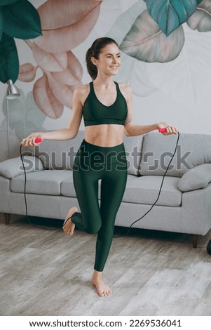 Full body young strong sporty athletic fitness trainer instructor woman wear green tracksuit jump use skipping rope look aside train do exercises at home gym indoor. Workout sport motivation concept