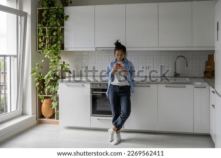 Full length of Asian woman housewife holding mobile phone using app to pay utility bills. Millennial girl standing in kitchen at home looking at smartphone screen searching cooking recipes Royalty-Free Stock Photo #2269562411