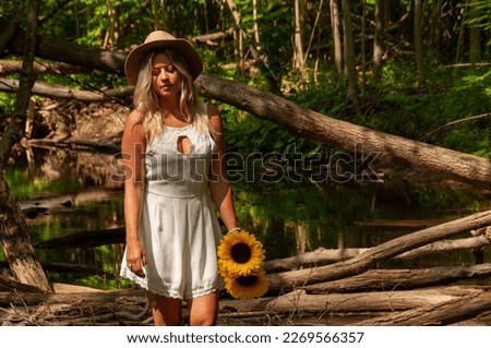 A beautiful young woman wearing a white dress and a hat holds sunflowers in a creek in the forest during a sunny summer's day