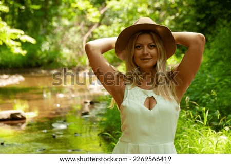 A young woman in a white dress wearing a hat has her arms behind her head on a sunny day in the forest