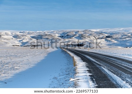 View along ring road route 1 in southern Iceland bending through a snowy mountainous landscape and road recently cleared of snow with sunlight on background against a clear blue sky